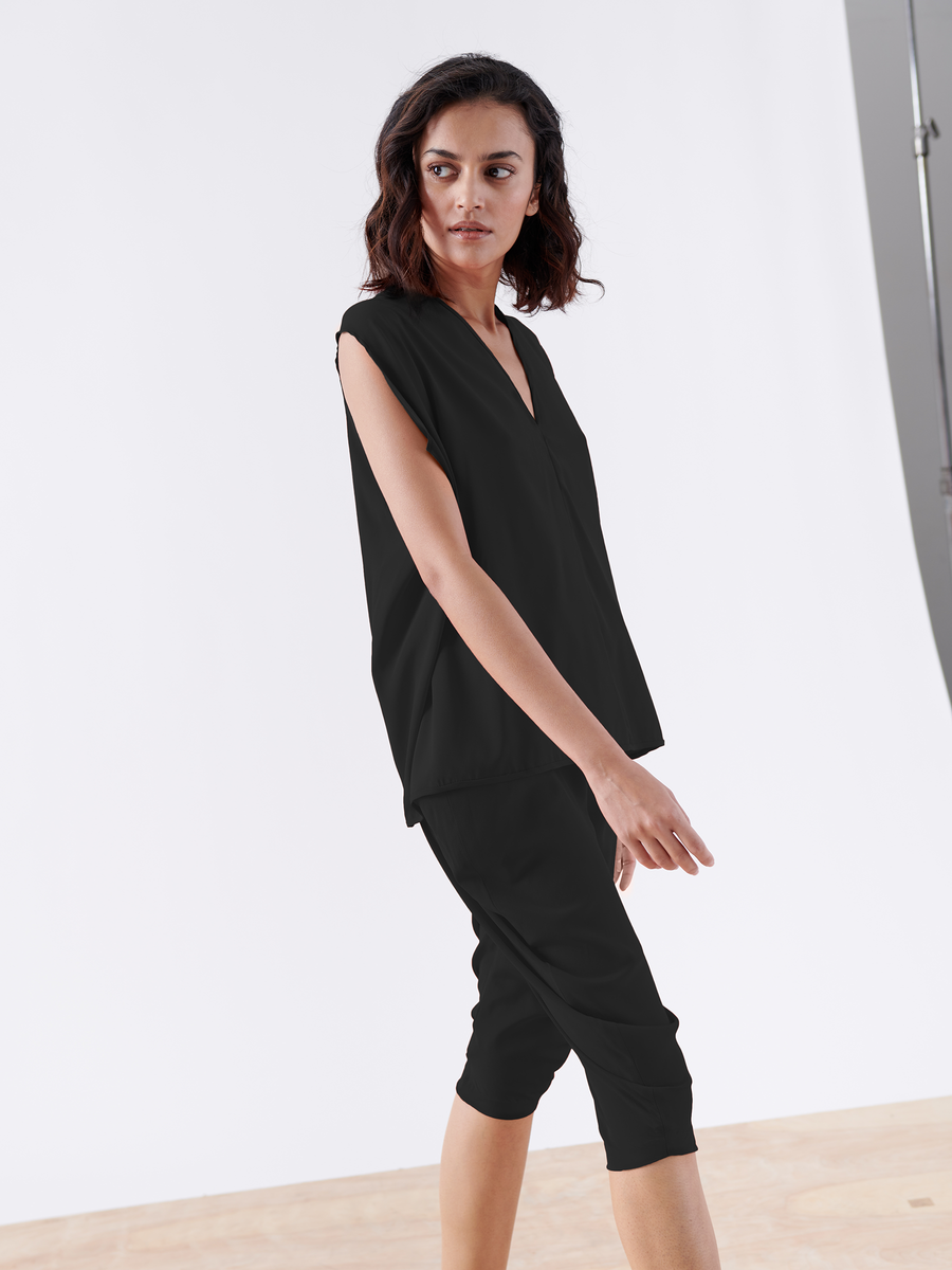 A person with medium-length dark hair walks on a wooden floor, wearing a sleeveless black top and the sleek Crop Curve Ori Pant by Zero + Maria Cornejo. The clear studio light and simple, light background evoke fashion made in New York.