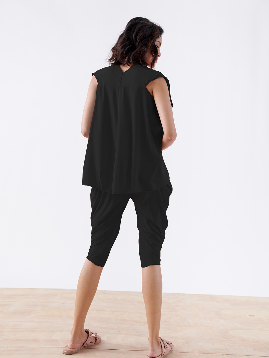 Someone wearing Zero + Maria Cornejo's black Crop Curve Ori Pant - Final Sale stands on a wooden floor, facing away. The pants feature an elastic banded waist and relaxed draping through the leg, paired with a sleeveless top and sandals against a plain white backdrop. Their wavy brown hair adds to the look.