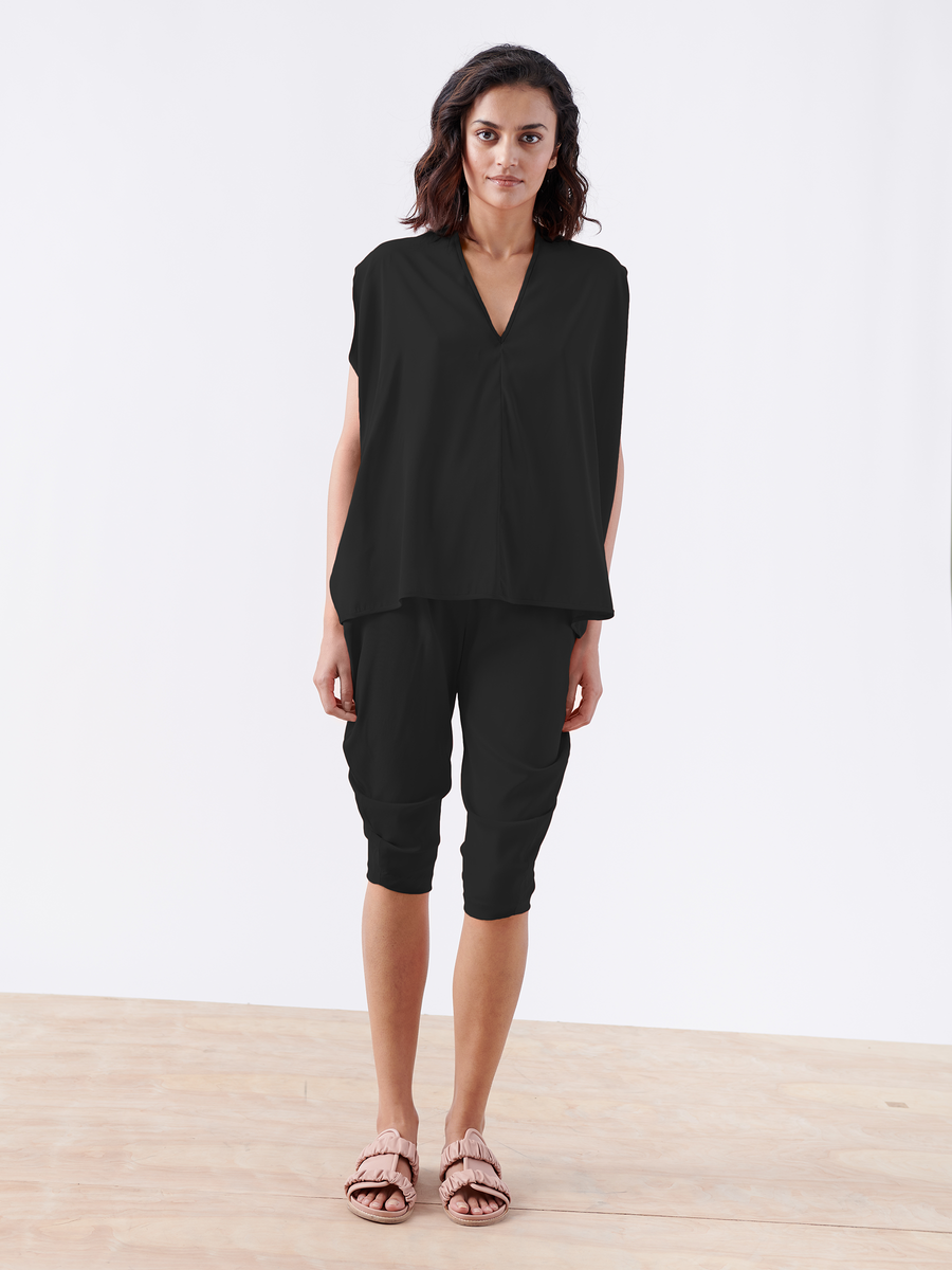 A woman with wavy shoulder-length hair stands on a light wooden floor against a white background, wearing the loose-fitting Crop Curve Ori Pant by Zero + Maria Cornejo, along with a loose black top and light pink sandals.