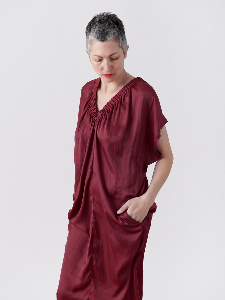 A person with short gray hair wears the Ruched Tilly Dress in deep red by Zero + Maria Cornejo. Against a light gray background, they thoughtfully look down, one hand in their pocket.