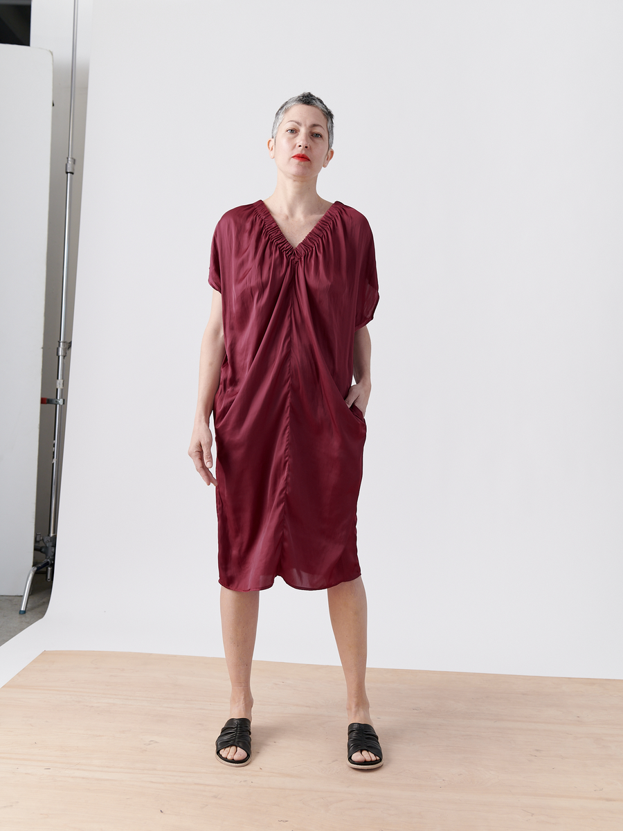 A confident person with short hair stands in a studio wearing the burgundy Ruched Tilly Dress by Zero + Maria Cornejo and black slide sandals. With hands in pockets and a neutral expression, they pose against a plain white background where part of a light stand is visible.