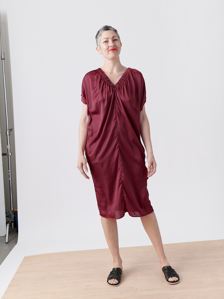 A person with short gray hair stands on a wooden platform against a plain white background, wearing the loose Ruched Tilly Dress in burgundy by Zero + Maria Cornejo, made of washable fabric. The knee-length v-neck dress complements their relaxed posture and slight smile, paired with black sandals.