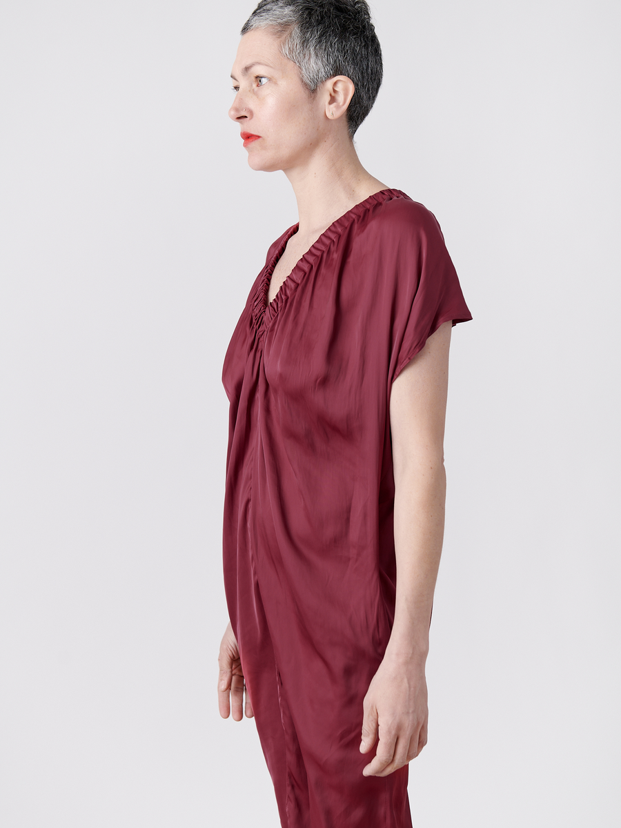 A person with short gray hair, wearing the Ruched Tilly Dress from Zero + Maria Cornejo in a loose-fitting burgundy v-neck style made of washable fabric, stands in profile facing left against a plain white background, with red lipstick and a calm expression.