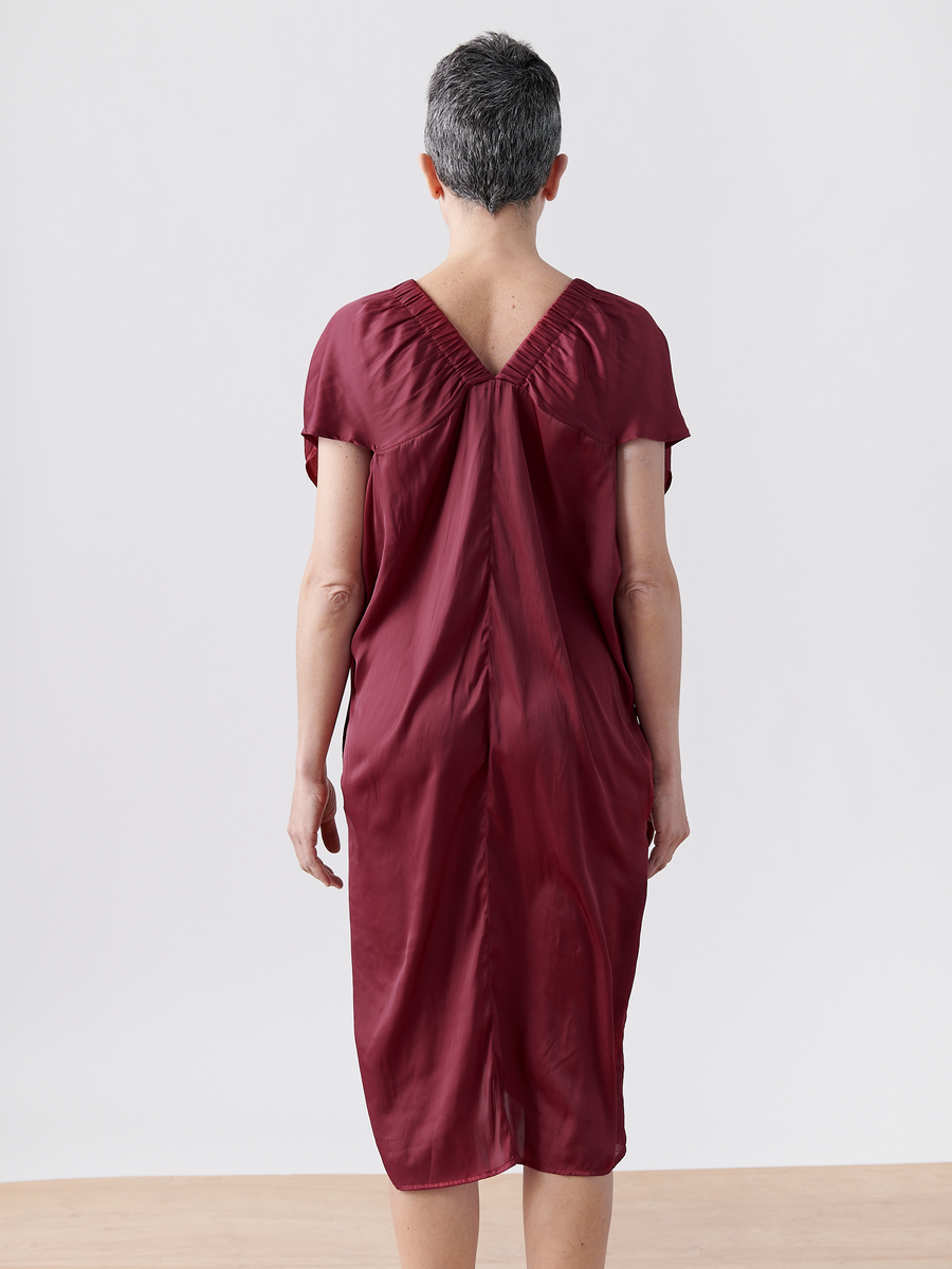 A person with short gray hair is wearing the Ruched Tilly Dress - Final Sale by Zero + Maria Cornejo. The knee-length, deep red silk v-neck dress features short sleeves. They stand on a wooden floor against a white background with their back facing the camera.