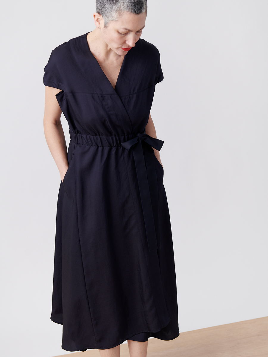 A short-haired, gray person in a dark Wrap Hex Dress by Zero + Maria Cornejo stands against a light background. The knee-length, short-sleeved dress is made from recycled viscose and features pockets where their hands are rested as they look down.