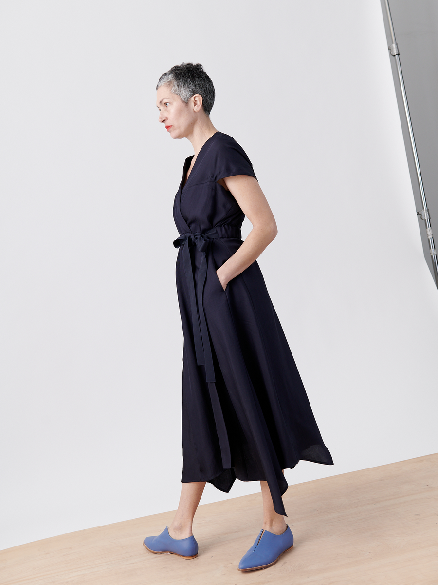 A woman with short gray hair models the Wrap Hex Dress by Zero + Maria Cornejo. This dark blue, sleeveless wrap dress features an asymmetrical hem and a belt, crafted from recycled viscose. She stands sideways with her hands in the pockets of the dress against a plain white background, wearing blue shoes.