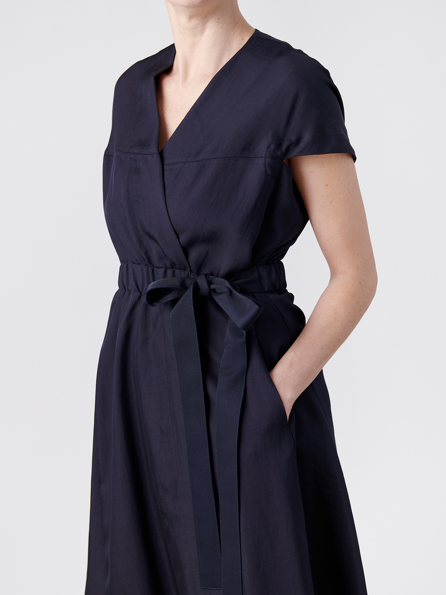 A person is wearing the Wrap Hex Dress - Final Sale by Zero + Maria Cornejo. This dark blue dress, made from recycled viscose, has short sleeves, a tie waist, v-neckline, pockets, and asymmetrical hem. Their head is partially out of the frame against a plain white background.