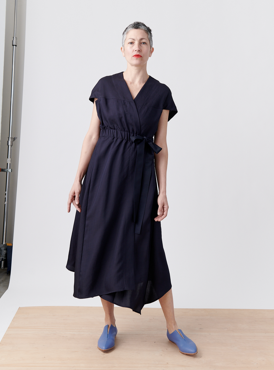 A person with short gray hair stands confidently on a wooden platform wearing the Wrap Hex Dress by Zero + Maria Cornejo, featuring an asymmetrical hem, paired with bright blue slip-on shoes. The backdrop is a plain light gray wall.