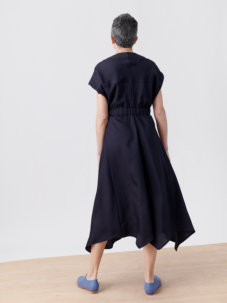 A person with short hair stands on a wooden floor, facing away from the camera, wearing Zero + Maria Cornejo's Wrap Hex Dress made from recycled viscose. The dress features a cinched waist and asymmetrical hem and is paired with light blue shoes against a plain white background.