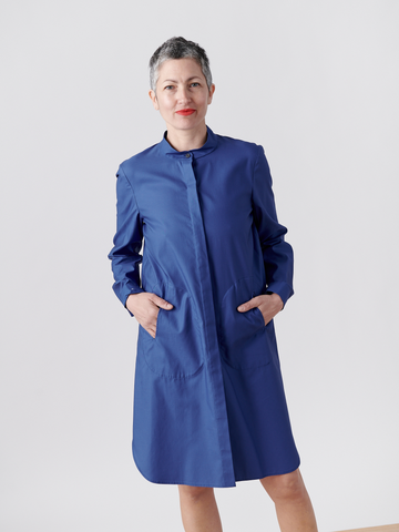 A woman with short gray hair smiles against a plain background in a Forward Shirt Dress by Zero + Maria Cornejo. The blue cotton broadcloth dress features a banded collar, hidden button-down placket, and pockets where she comfortably tucks her hands.