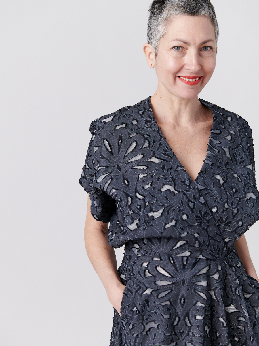 A person with short gray hair smiles, wearing the Aki Wave Dress by Zero + Maria Cornejo. This dark, floral-patterned dress features a wrap-effect V-neck and short sleeves. The background is plain and light-colored.