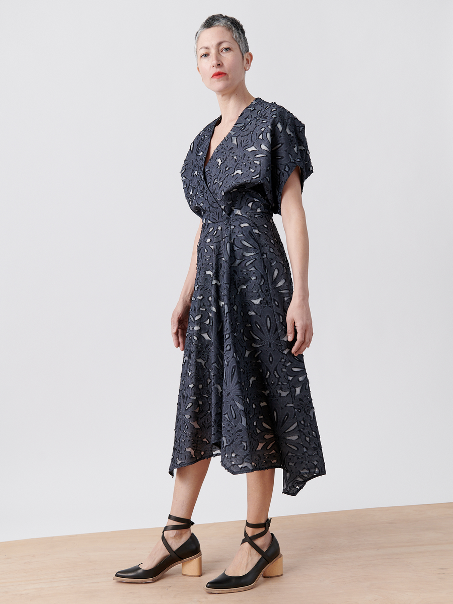 Someone in an Aki Wave Dress by Zero + Maria Cornejo, a dark blue wrap-effect v-neck dress, stands on wood against a white background. With short hair and black ankle-strap heels, they gaze slightly upward and to the side, exuding elegance.