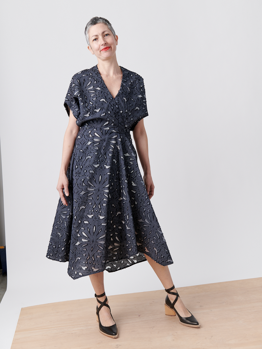 Sporting a confident stance, a person with short hair showcases the navy blue Aki Wave Dress from Zero + Maria Cornejo. Made from organic cotton yarns, it features a floral-patterned wrap-effect V-neck. Stylish ankle-strap block heel shoes complete their look against a neutral background.