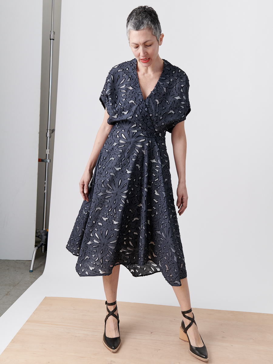 A person with short gray hair is wearing the Zero + Maria Cornejo Aki Wave Dress, a floral-patterned wrap-effect v-neck dress, and black strappy shoes. They stand on a wooden platform in a studio with white and gray backgrounds, looking down at their feet.