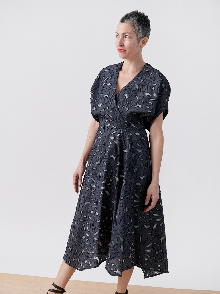 A person with short hair is wearing the Aki Wave Dress by Zero + Maria Cornejo, featuring luxurious damask lace jacquard and floral cutouts. They stand against a plain white background, looking to the side with a relaxed expression, complemented by black shoes.