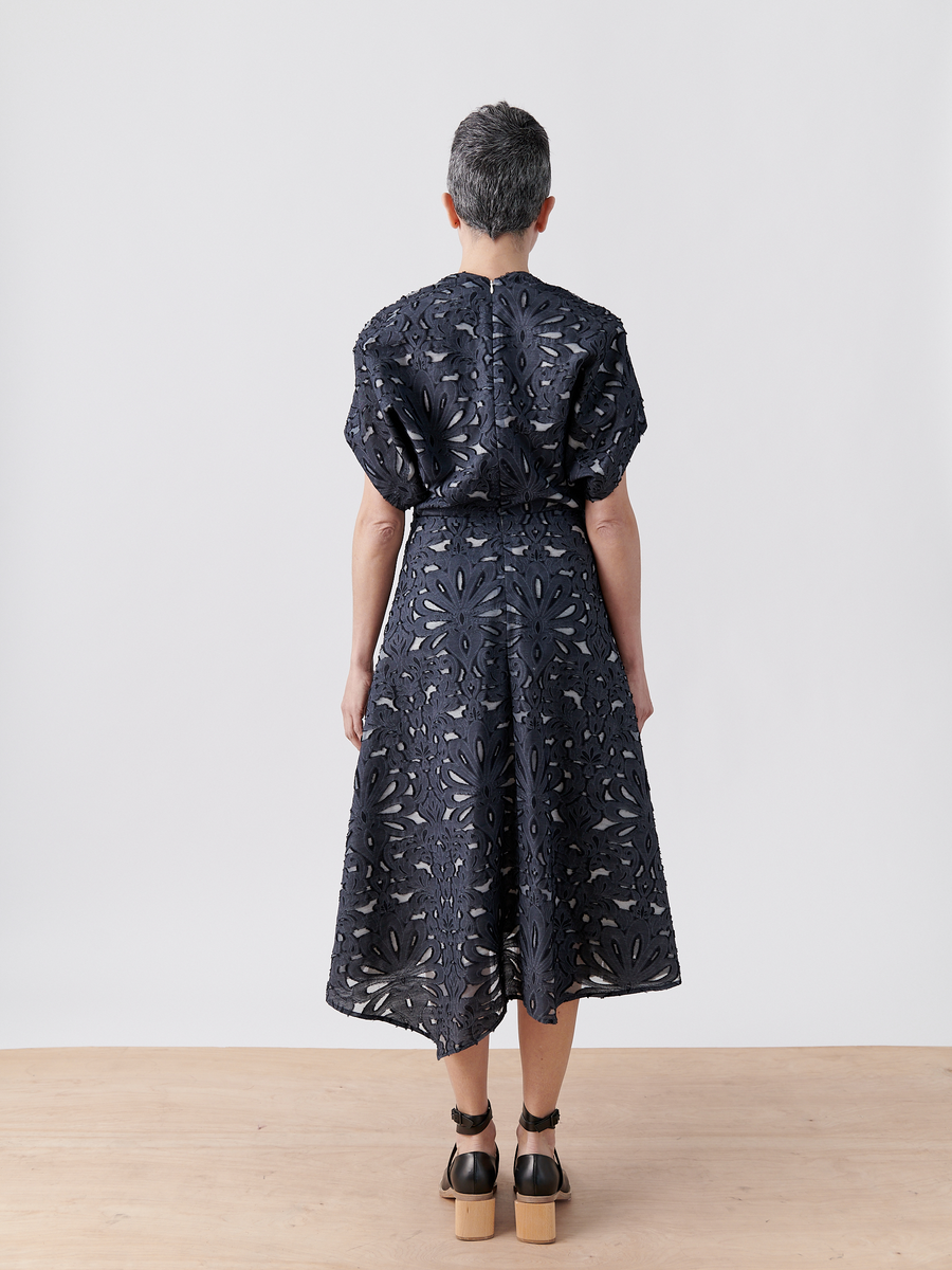 A person with short gray hair wears the Aki Wave Dress by Zero + Maria Cornejo, a navy floral-patterned wrap-effect V-neck dress. Standing on a wooden floor against a light gray background, they complete the look with black platform shoes made from organic cotton yarns.