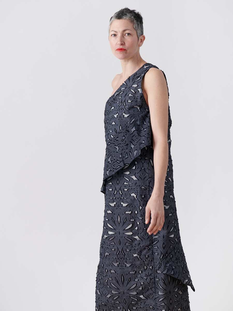 A person with short gray hair is wearing a sleeveless dark sheath dress called the Long Spiral Dress, featuring intricate Damask lace jacquard, by Zero + Maria Cornejo. They stand against a plain white background, looking forward with a neutral expression.