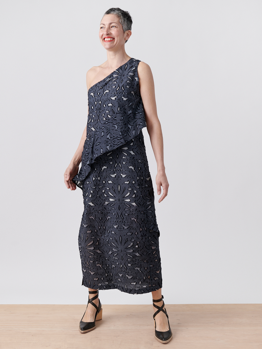 A person stands against a plain background, smiling in the Zero + Maria Cornejo Long Spiral Dress, a navy blue one-shoulder sheath with floral lace details. Black strappy shoes complete the ensemble, offering a perfect blend of elegance and style.