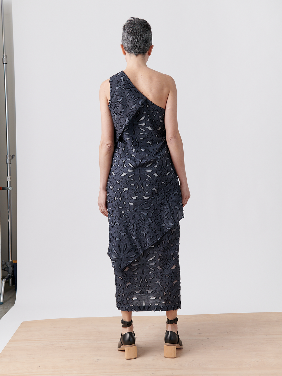 A person with short gray hair stands on a wooden floor, facing away, wearing Zero + Maria Cornejo's Long Spiral Dress in navy Damask lace jacquard with floral patterns and platform sandals against a plain white background.