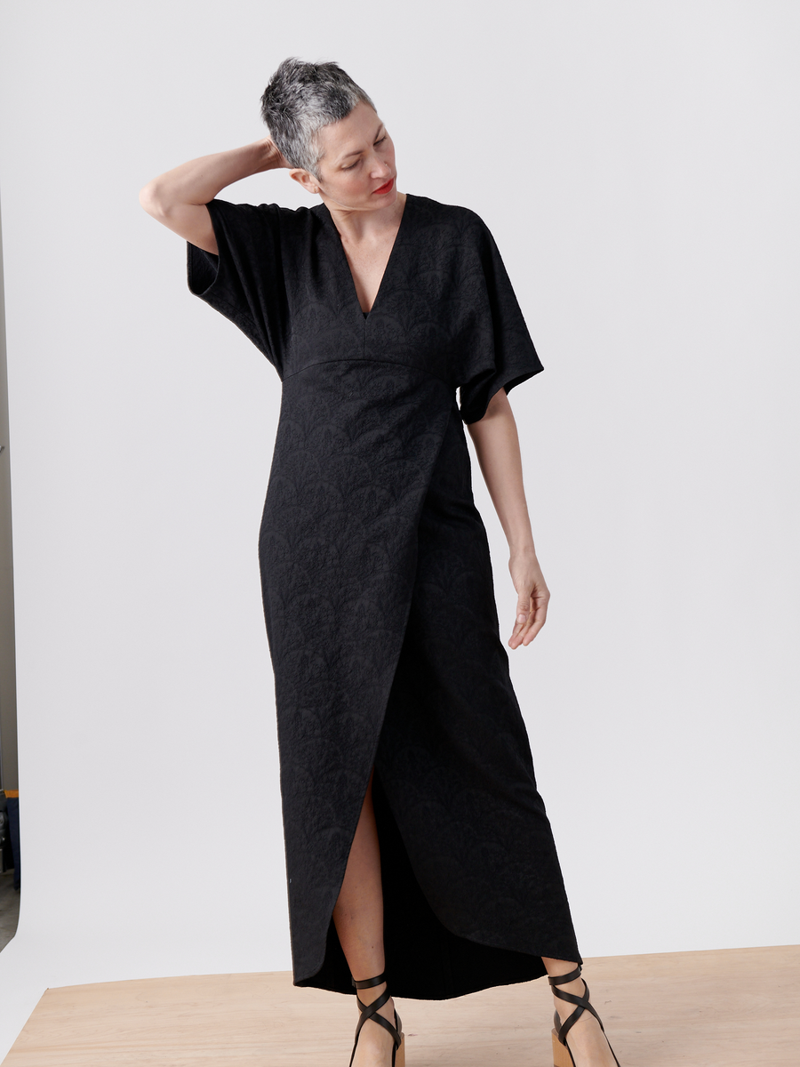 A woman with short hair models the Zero + Maria Cornejo Wrap Eri Dress, a long, dark stretch jacquard dress with a subtle floral pattern. On a wooden floor and light gray background, she looks down, one hand behind her head, and wears strappy high heels.