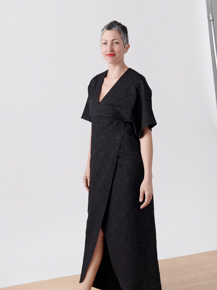 A confident person with short gray hair smiles while wearing the Wrap Eri Dress by Zero + Maria Cornejo. The dress, elegant with a deep V-neck, short sleeves, floral jacquard pattern, and front slit, contrasts against a light-colored floor and plain backdrop.