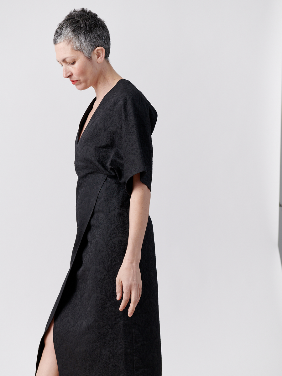 A serene individual with short gray hair and relaxed arms stands in profile against a plain white background, wearing the Wrap Eri Dress - Final Sale by Zero + Maria Cornejo, a black stretch jacquard dress featuring a side slit.