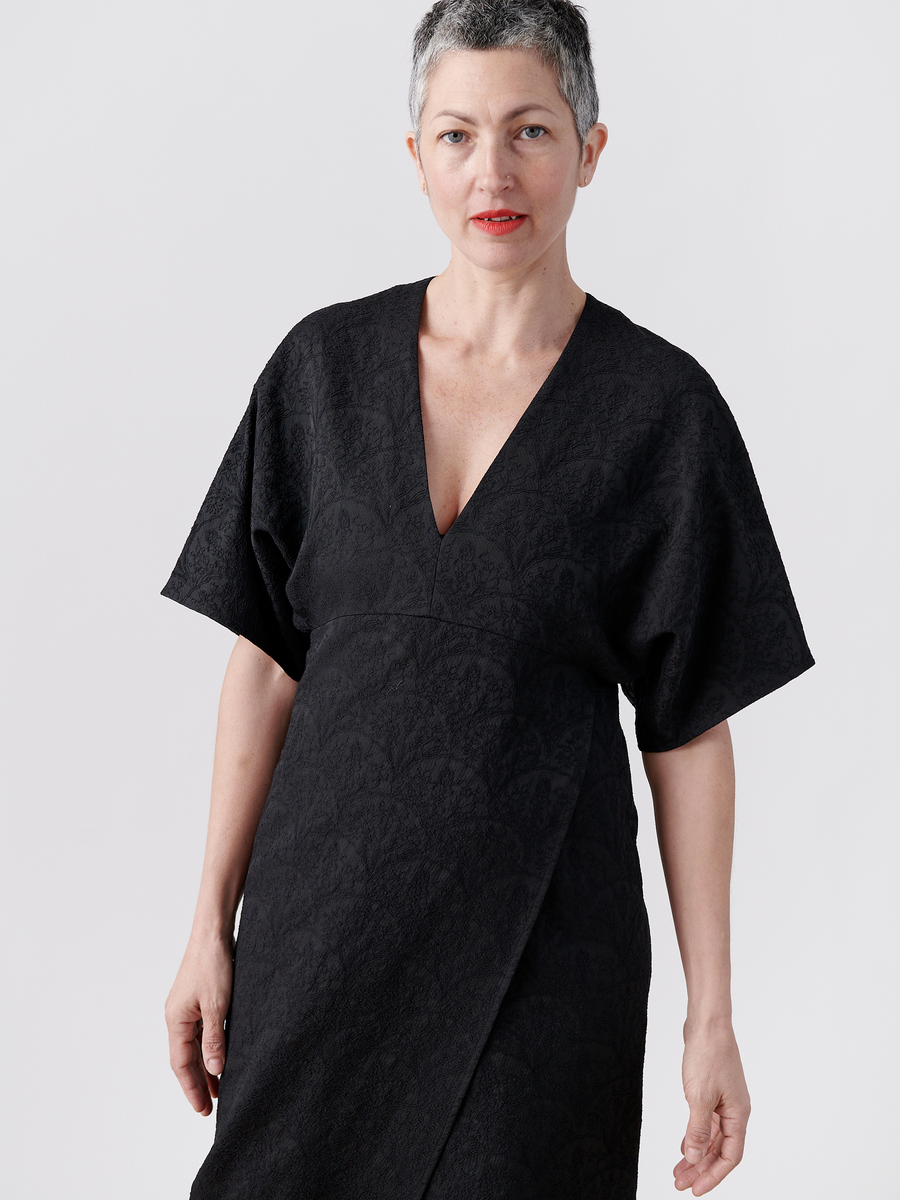 A person with short gray hair is wearing a Wrap Eri Dress from Zero + Maria Cornejo. The black V-neck stretch jacquard dress features loose sleeves and a subtle floral pattern, made from recycled polyester. They stand against a plain white background, looking into the camera with a slight smile.