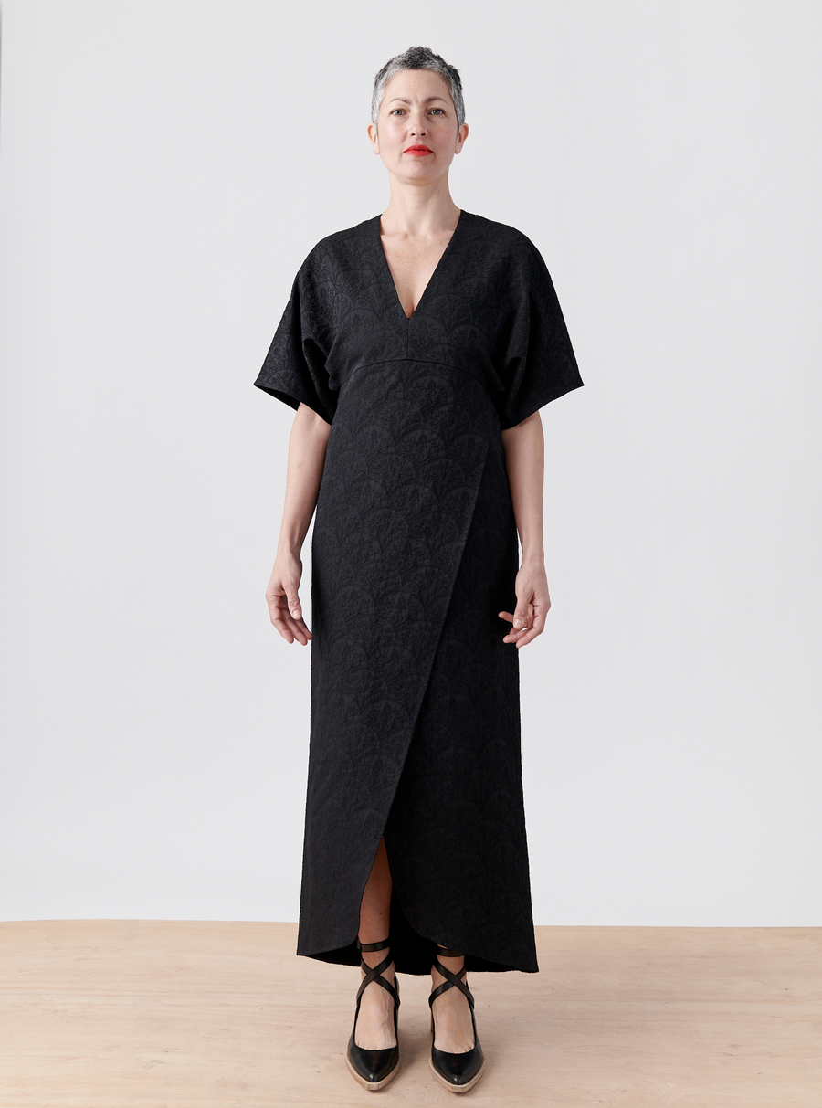 A person with short gray hair stands on a light wood floor wearing Zero + Maria Cornejo's black Wrap Eri Dress, made from recycled polyester, featuring short sleeves and paired with black ballet-style shoes. The background is plain white.