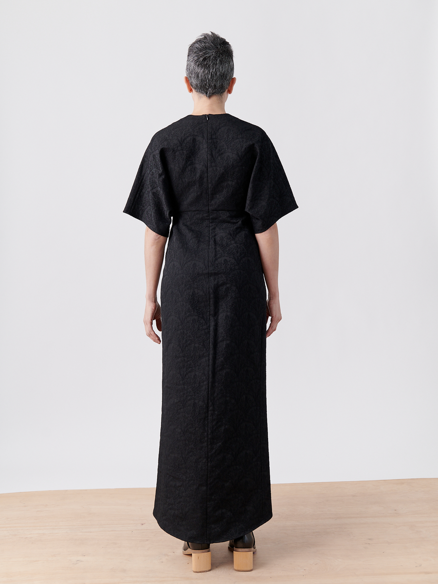 A person with short gray hair stands on a wooden floor, facing away, wearing the Wrap Eri Dress - Final Sale by Zero + Maria Cornejo. Its sleek design contrasts against the plain white background while paired with high platform shoes.