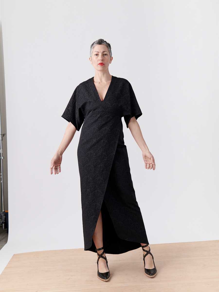 Against a plain backdrop, a person with short hair stands on a wooden platform wearing the Wrap Eri Dress by Zero + Maria Cornejo. The black dress in recycled polyester has short sleeves and a high slit. They wear red lipstick and black strapped shoes.