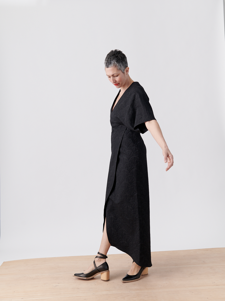 A person with short gray hair wears the Wrap Eri Dress - Final Sale by Zero + Maria Cornejo and black shoes with wooden block heels. They stand on a wooden floor, slightly turned to the side against a plain white background.