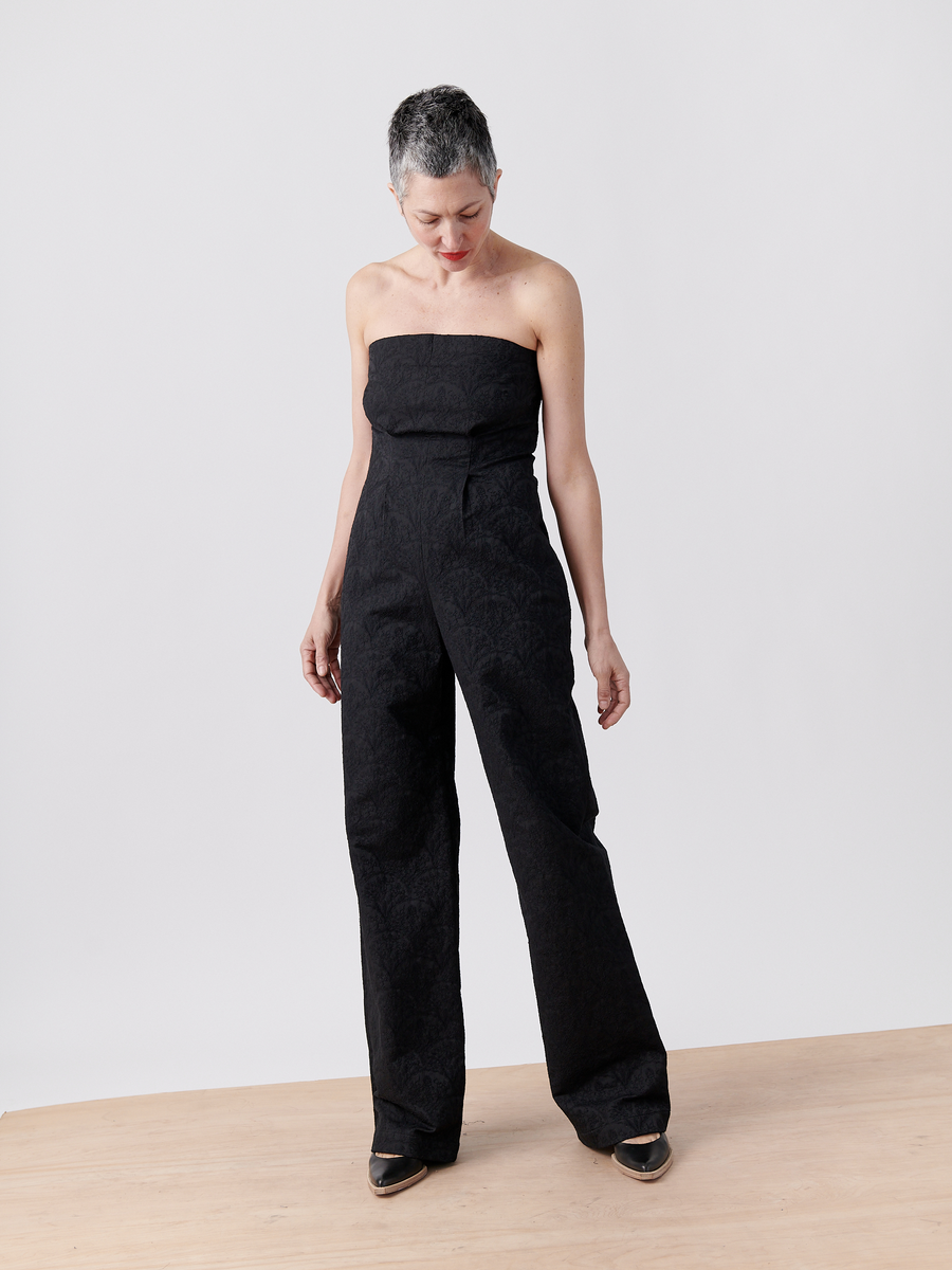 A person with short gray hair is modeling the Pia Jumpsuit by Zero + Maria Cornejo, crafted from sleek jacquard fabric, paired with black heels. They stand on a wooden surface against a plain white background, gazing down to their right.