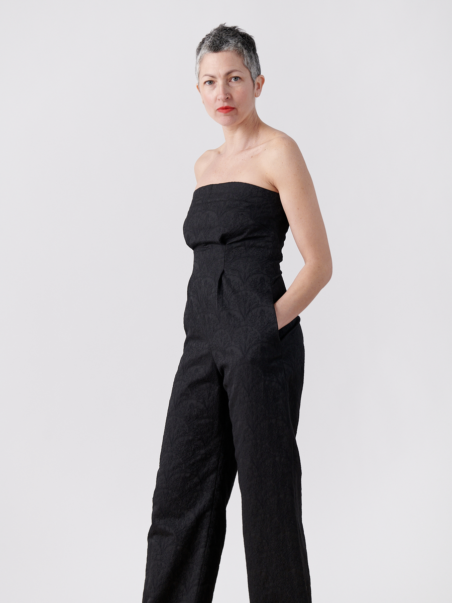 A person with short gray hair models the Pia Jumpsuit by Zero + Maria Cornejo in a simple black jacquard style. Standing against a plain white background, their confident posture, hands in pockets, and direct gaze create a striking image.