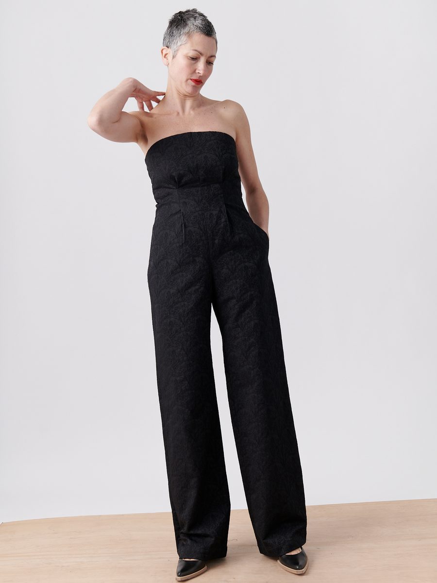 A person with short gray hair poses in the Zero + Maria Cornejo Pia Jumpsuit - Final Sale. The strapless black jumpsuit is made from recycled polyester, features pockets, and is paired with black pointed shoes on a wooden surface against a white background.