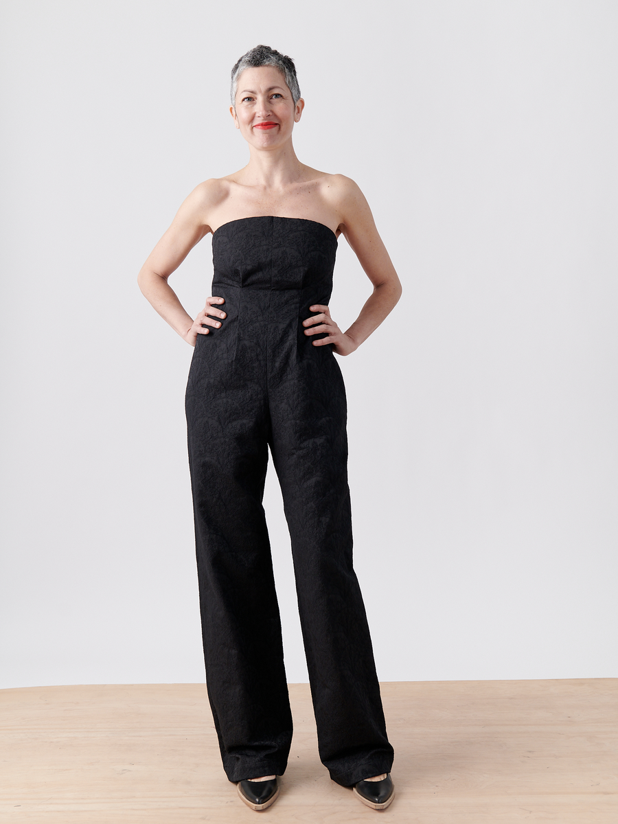 A person with short gray hair is wearing the Pia Jumpsuit by Zero + Maria Cornejo, a strapless black jumpsuit made from recycled polyester. They pair it with black shoes and stand smiling with hands on hips against a plain white background.