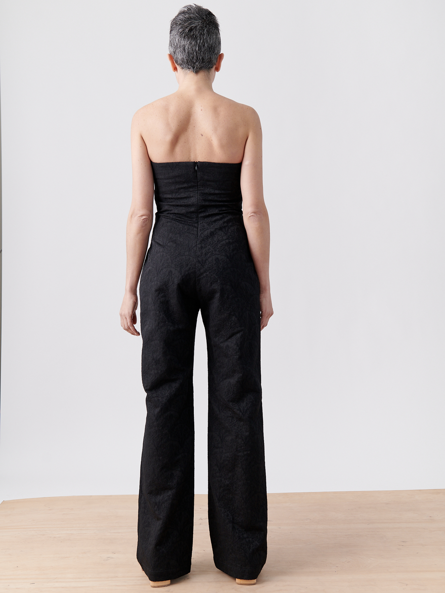 A person with short gray hair is seen from the back, wearing a strapless Pia Jumpsuit from Zero + Maria Cornejo. It's made of recycled polyester and they stand on a light wooden floor against a plain white background.