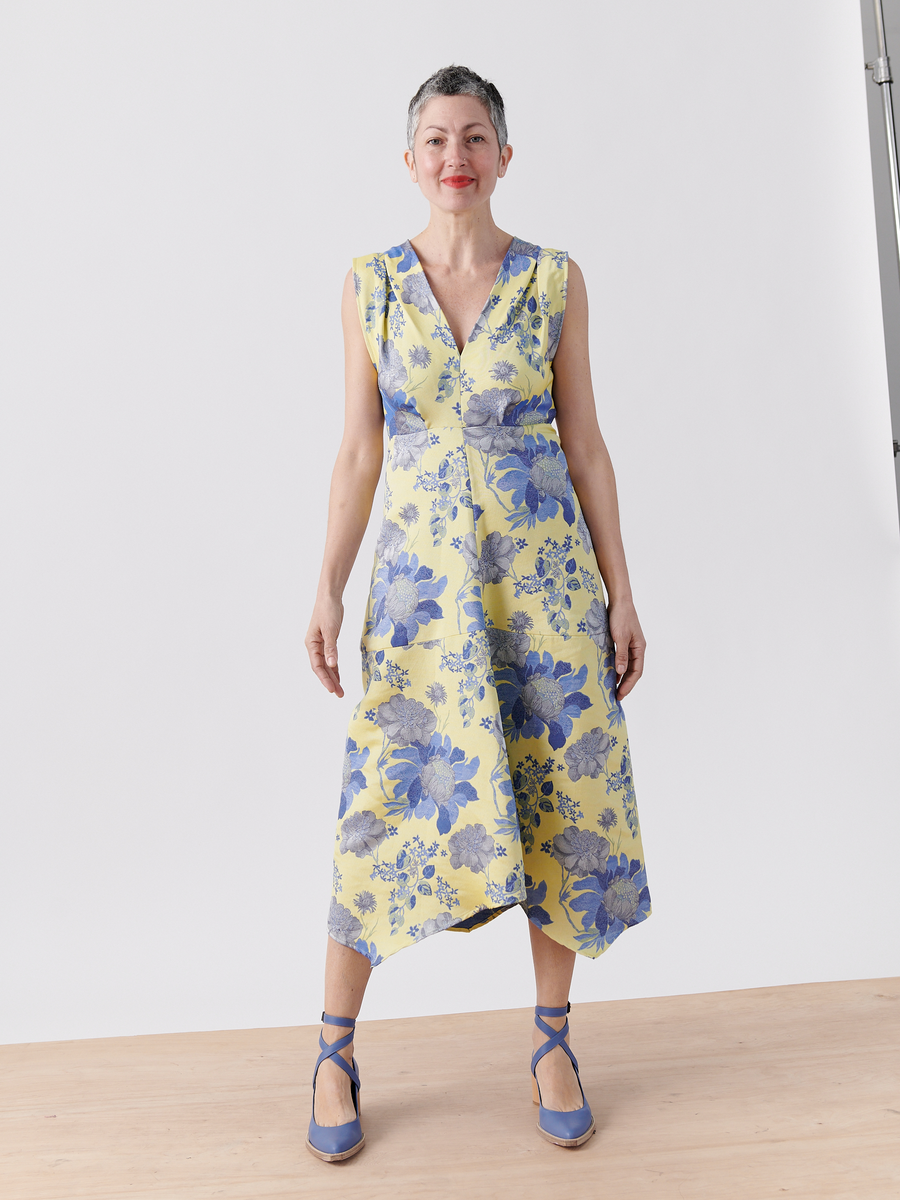 A person stands confidently in the Zero + Maria Cornejo Joi Dress - Final Sale, a sleeveless yellow floral jacquard dress with a V-neck and blue floral pattern. They pair it with blue ankle-strap heels against a plain white wall and light wooden floor, perfect for highlighting this stunning cocktail dress.