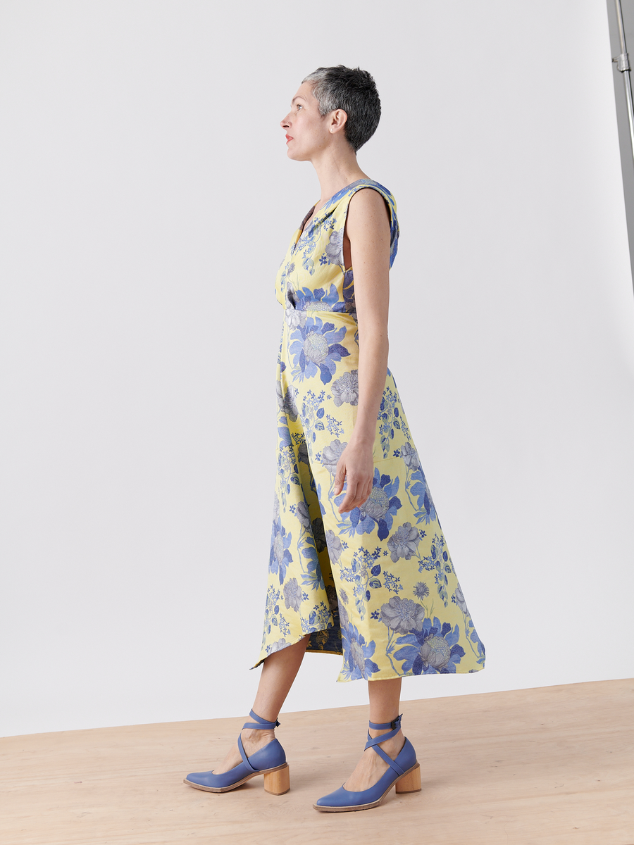 Side view of a person with short hair wearing the Joi Dress - Final Sale by Zero + Maria Cornejo, a sleeveless yellow floral jacquard design. They're in blue ankle-strap heels on a wooden floor against a white background.