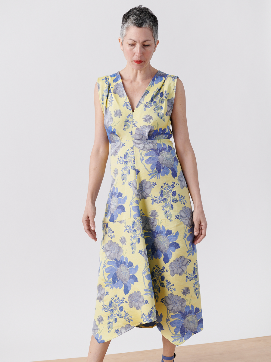 A person with short gray hair is wearing the Joi Dress from Zero + Maria Cornejo, a sleeveless yellow floral jacquard dress with blue patterns. It features a V-neck and flows to mid-calf length, set against a light floor and white background. Made of organic cotton, it’s on final sale.