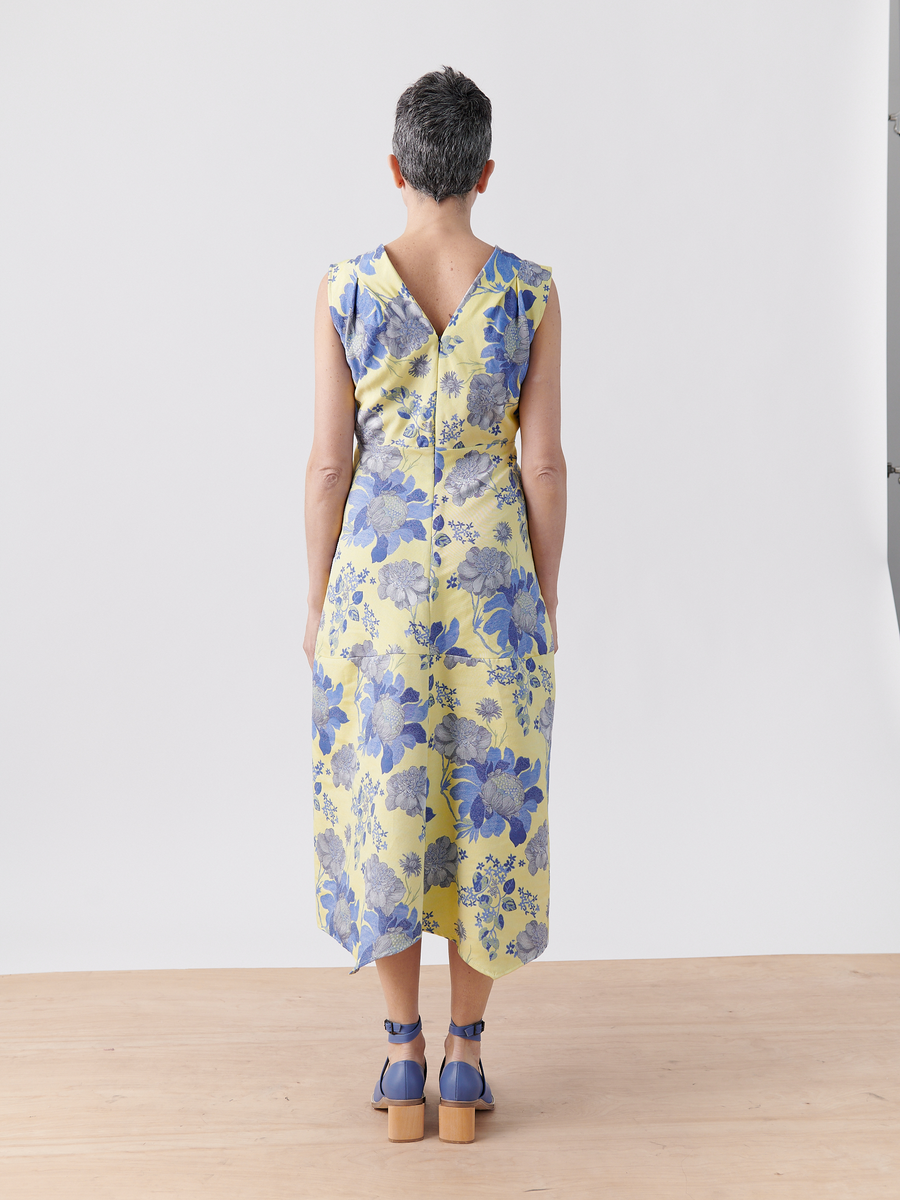 A person with short hair stands on a wooden floor, facing away, wearing the Joi Dress in yellow and blue patterns by Zero + Maria Cornejo paired with matching blue block-heeled shoes. The pristine white background adds elegance to the scene.