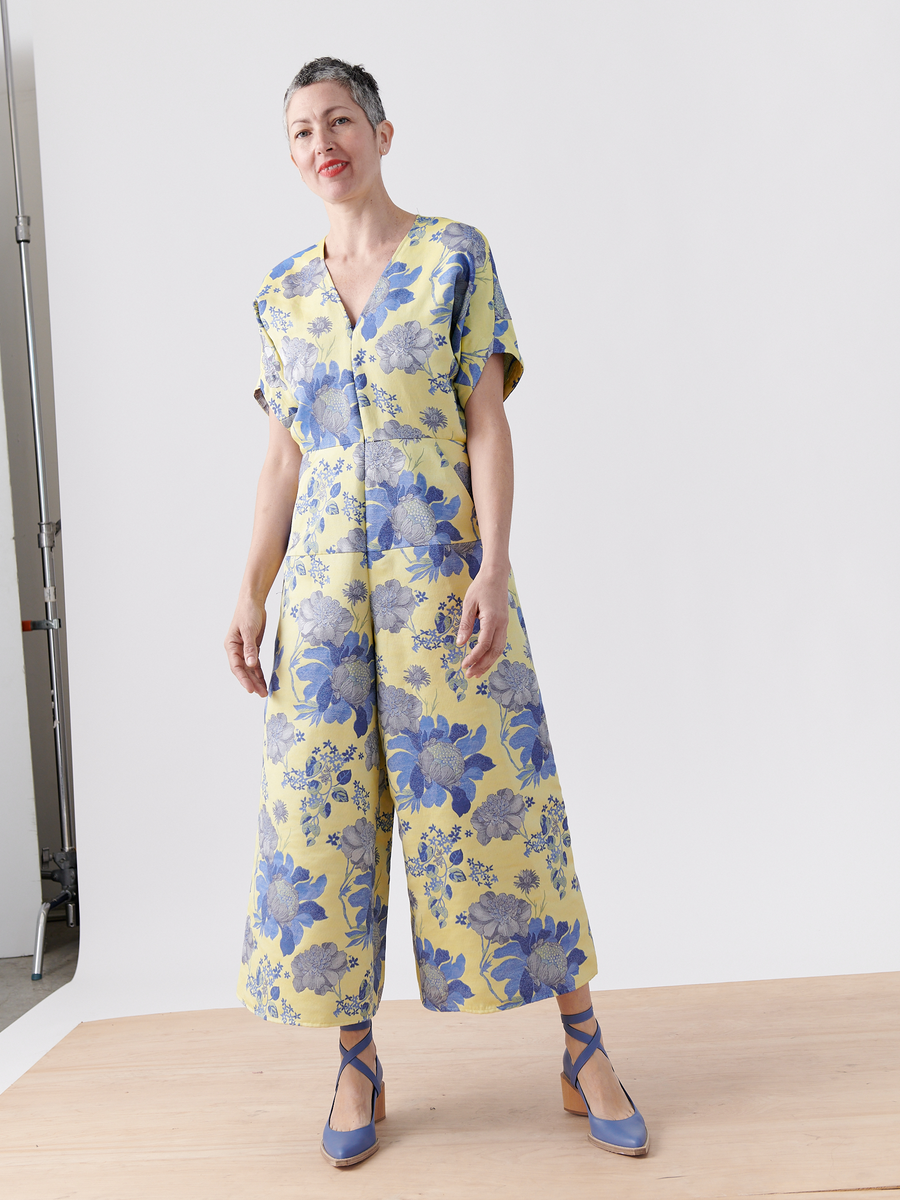 A person models the Aissa Eve Jumpsuit from Zero + Maria Cornejo, featuring large blue floral patterns on organic cotton. They have short gray hair and wear blue strappy shoes. The cropped wide-leg design harmonizes with the plain, light-colored studio setting beautifully.