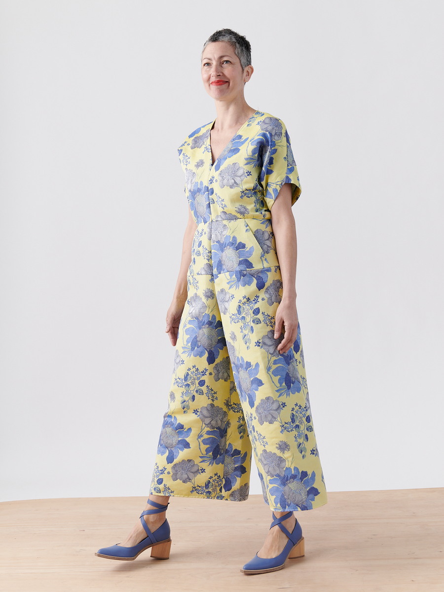 A person with short hair smiles, standing on a wooden floor, wearing the vibrant Aissa Eve Jumpsuit from Zero + Maria Cornejo. Made of organic cotton in a yellow floral jacquard design, it's paired with blue heels. The cropped wide-leg style stands out against the plain, light background.