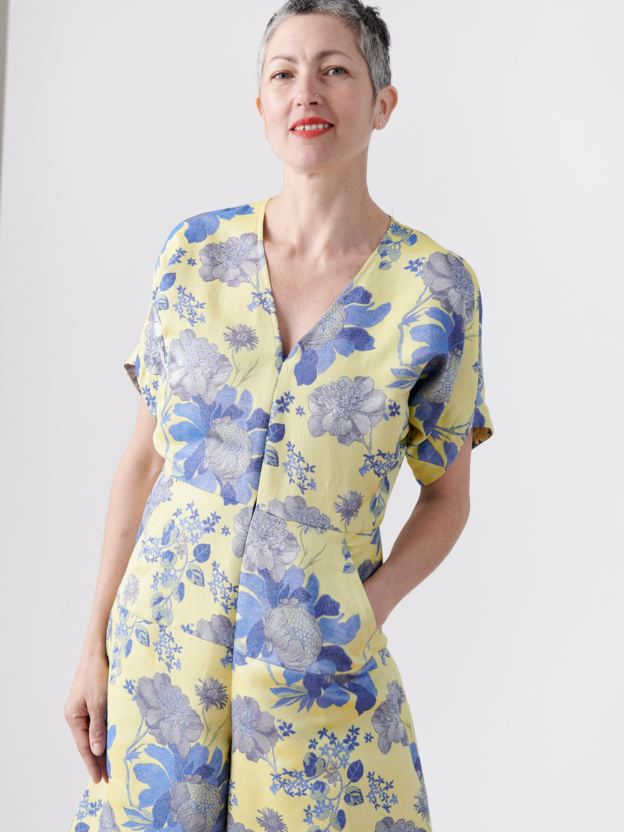 A smiling person with short hair poses against a white background in the Aissa Eve Jumpsuit by Zero + Maria Cornejo. The yellow floral jacquard, organic cotton jumpsuit boasts a deep V-neckline, short sleeves, and pockets for an effortlessly stylish look.