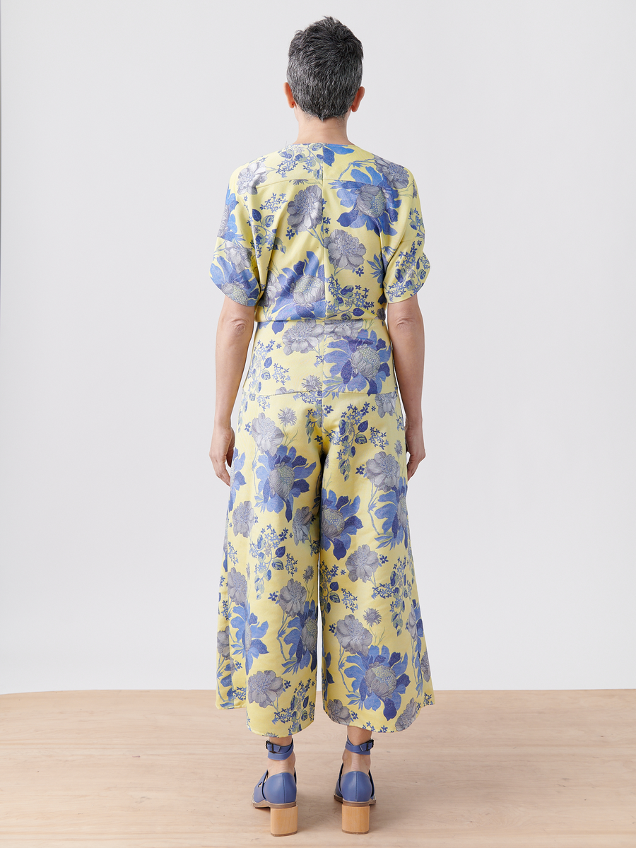 A person with short hair faces away, wearing the Aissa Eve Jumpsuit by Zero + Maria Cornejo—a vibrant floral jacquard design with cropped wide-leg pants—and blue platform shoes on a light wooden floor against a plain backdrop.