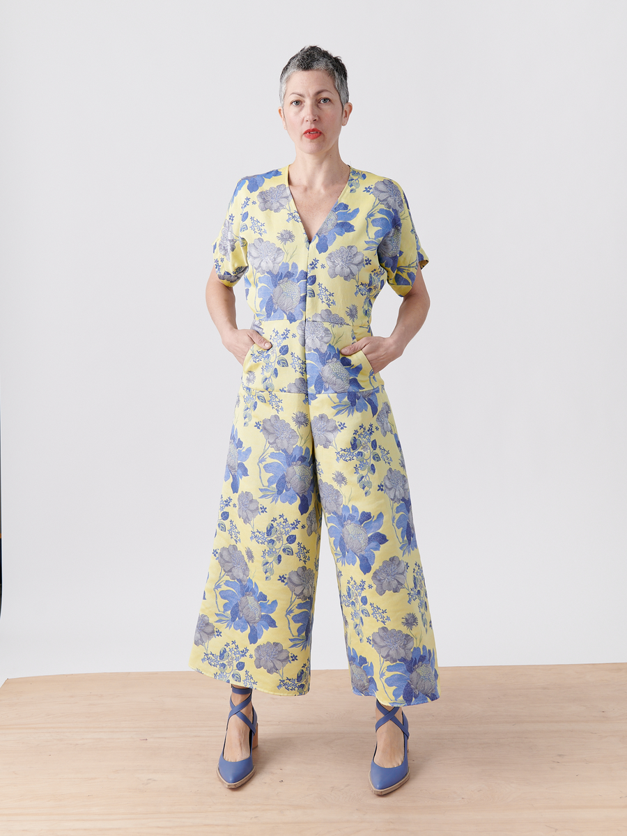 A person confidently stands on a wooden floor, wearing the Aissa Eve Jumpsuit by Zero + Maria Cornejo in yellow floral jacquard made of organic cotton. Their hands are in their pockets, and they don blue shoes with crisscross straps against a plain white background.