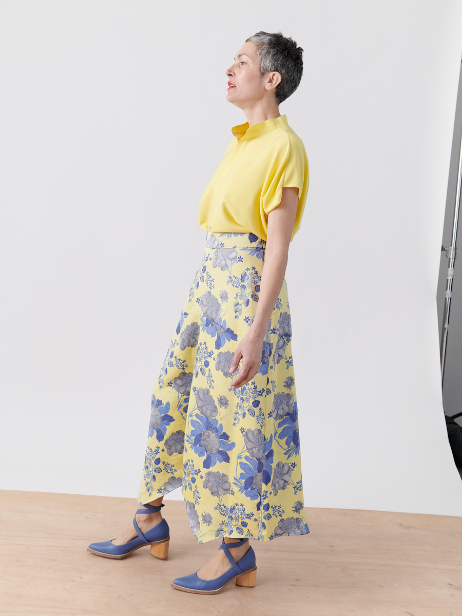 A person with short hair is on a wooden floor, wearing a yellow, short-sleeve blouse paired with the Joi Skirt from Zero + Maria Cornejo. The high-waist skirt features blue floral jacquard patterns. They wear blue heels and glance upwards to the side against a plain white background.