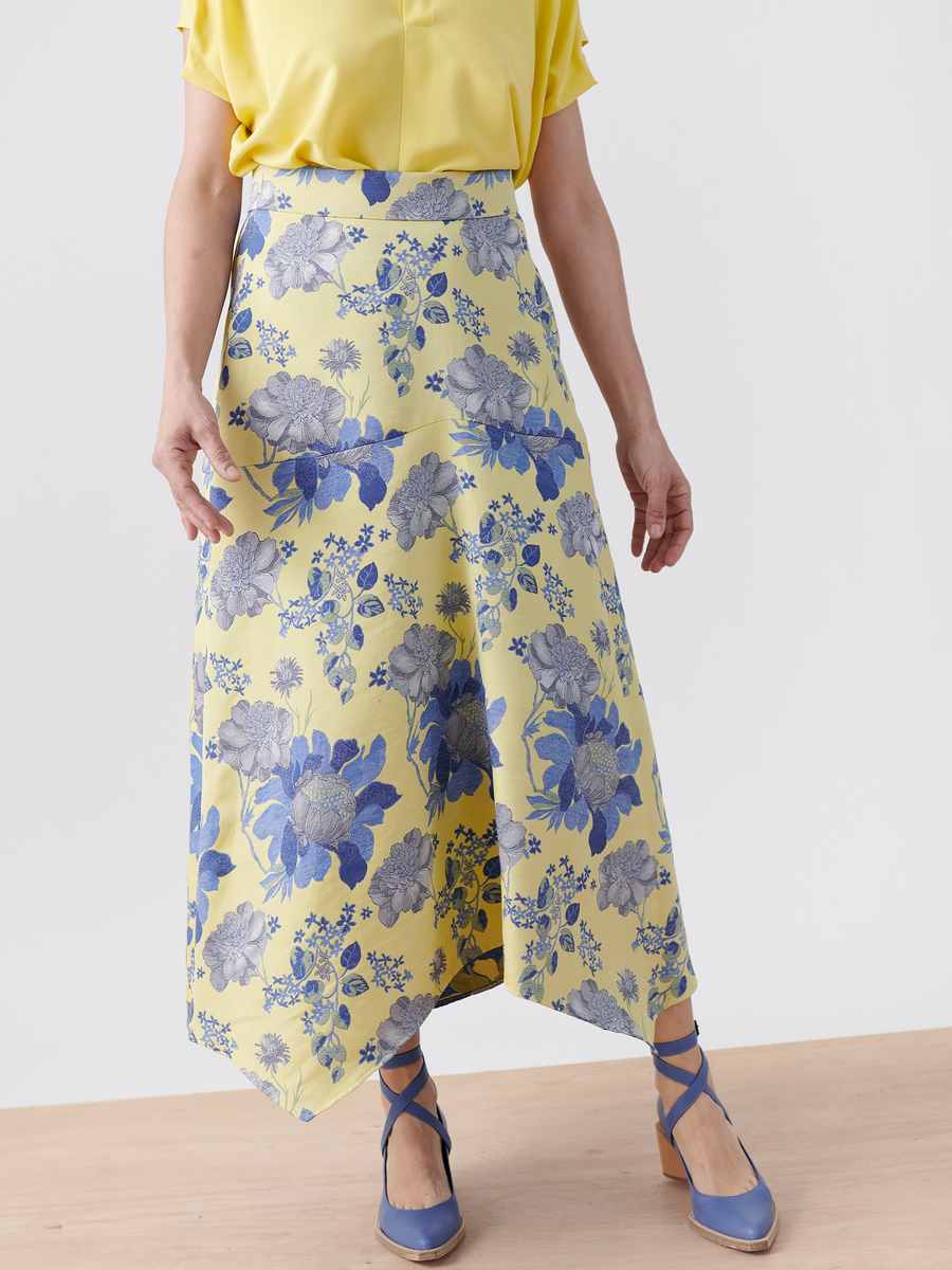 A person showcases the Joi Skirt - Final Sale from Zero + Maria Cornejo, paired with a yellow blouse and blue ankle-strap heels, on a light wooden floor. The image spotlights the skirt's intricate details from the waist down.