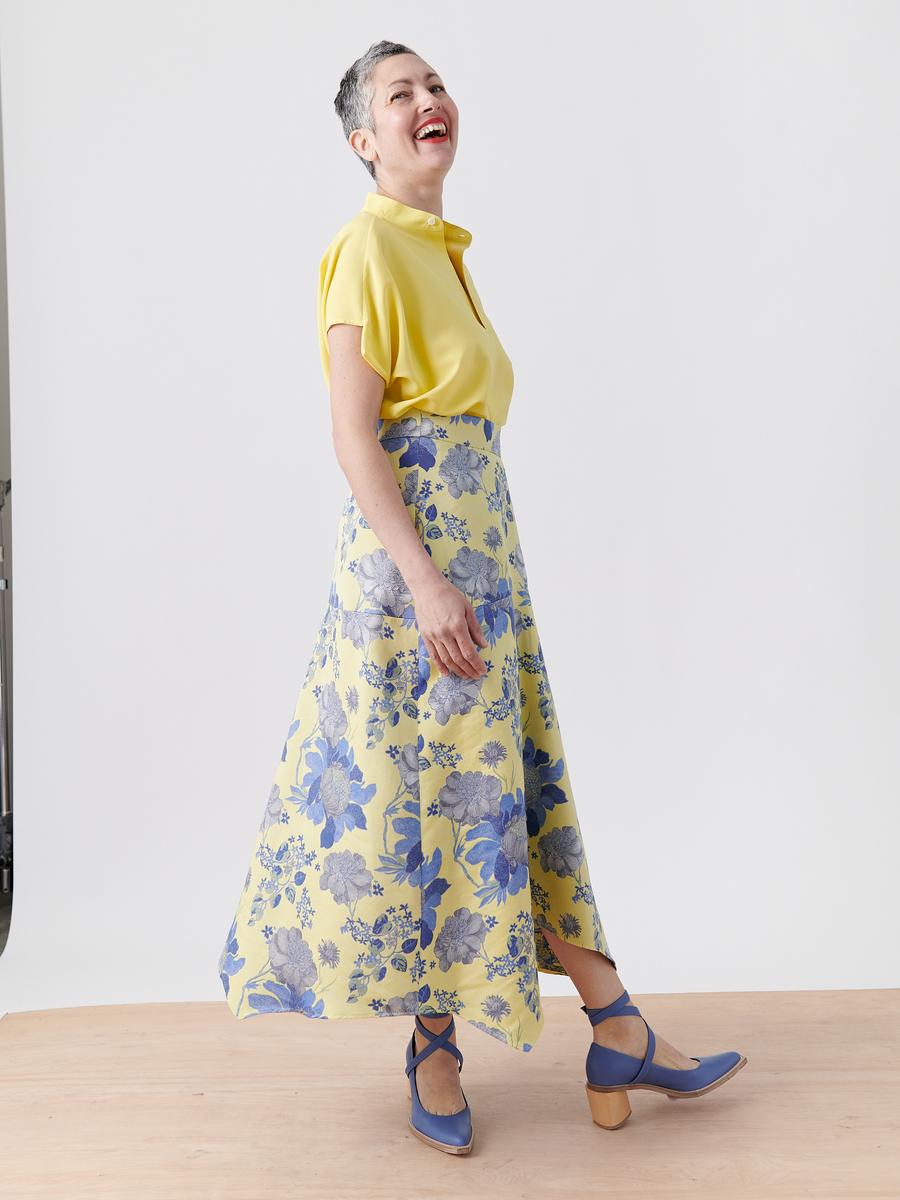 A woman with short gray hair wears a yellow blouse and the Joi Skirt - Final Sale by Zero + Maria Cornejo, paired with blue heels. She stands on a light wooden floor against a plain white background, smiling and looking upward.