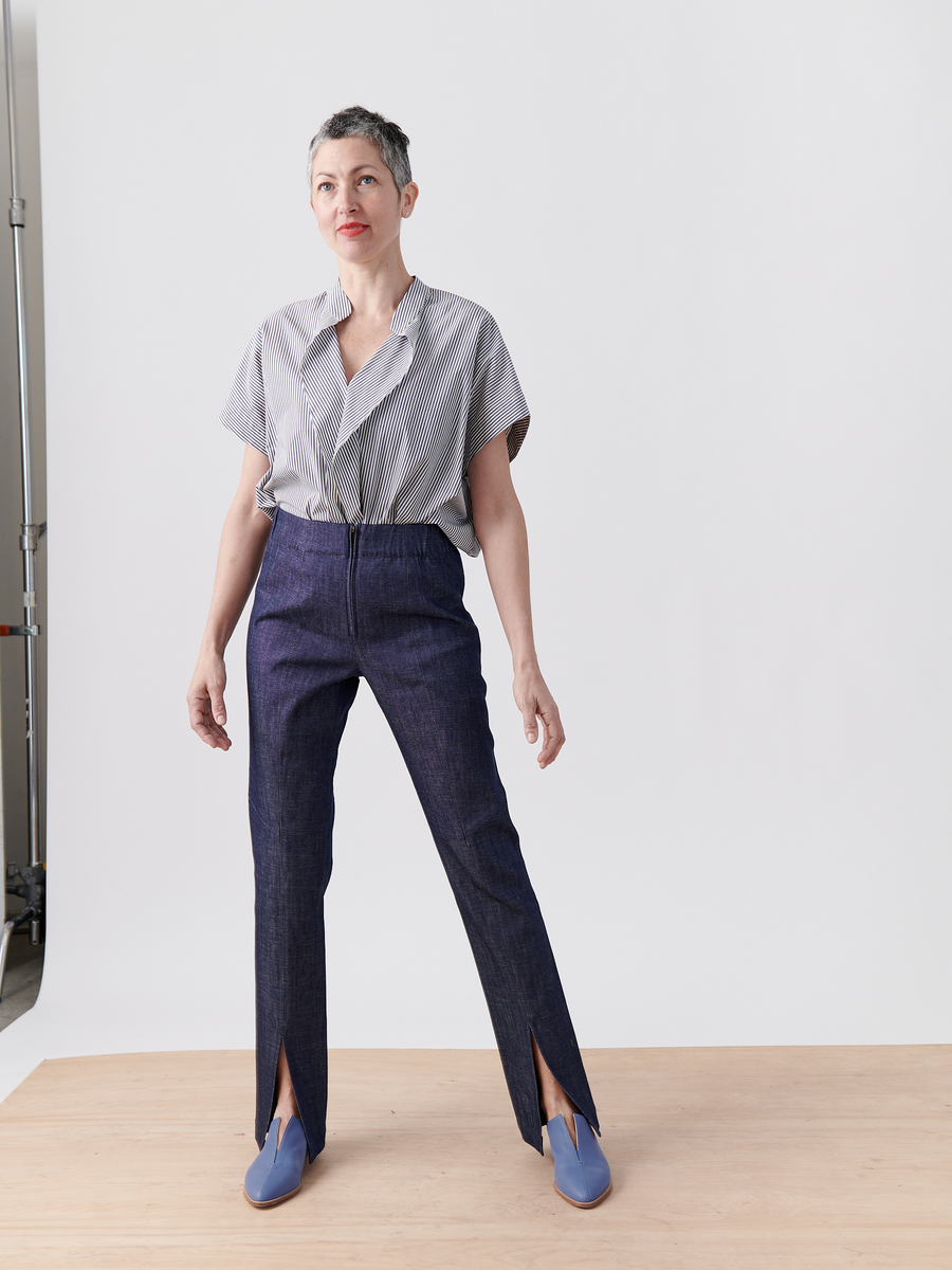 A person with short grey hair confidently stands on a wooden surface in a blue-and-white striped shirt and wearing Zero + Maria Cornejo's Denim Tala Trouser, made of 100% organic cotton with front slits, paired with blue shoes, set against a plain white background.