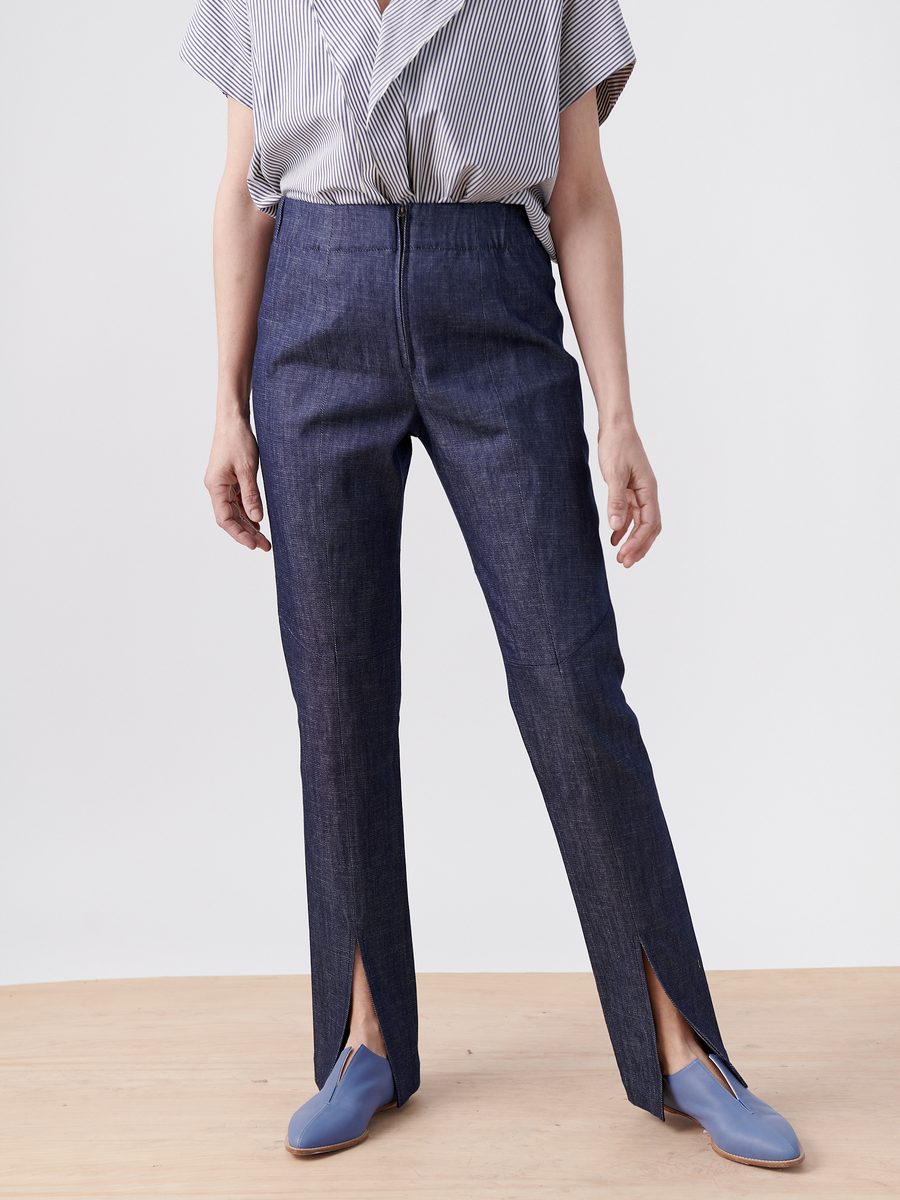 A person in high-waisted Denim Tala Trousers by Zero + Maria Cornejo, Cradle 2 Cradle Certified with front slits, a striped short-sleeve blouse, and blue slip-on shoes stands on a light wood floor against a plain background.